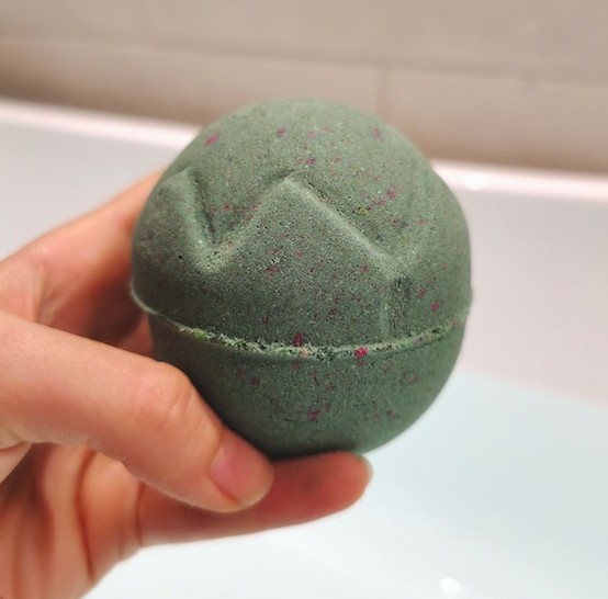 hand holding Lord of Misrule Bath Bomb by Lush in front of a bath